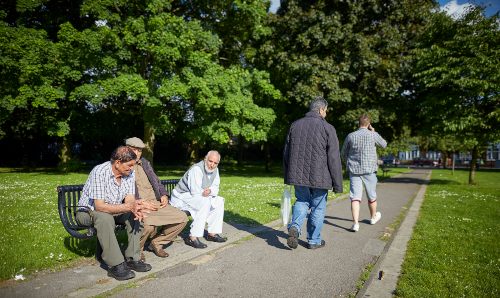 Two men sat on a bench in a park, while two other men walk past.