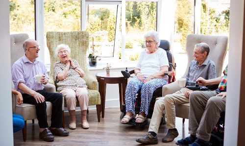 A group of elderly male and female people sat in a care home talking.