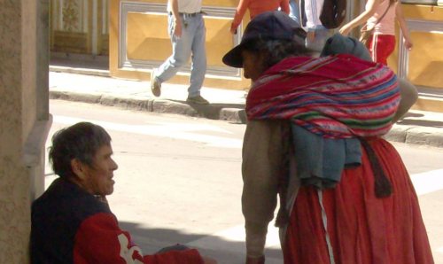 An older lady talking to an older man sat on the street floor. 